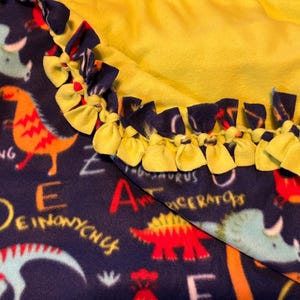 May include: A navy blue fleece blanket featuring colorful dinosaur illustrations and text. The blanket has a bright yellow backing and a knotted edge in yellow and navy blue. The dinosaurs are in shades of orange, red, and teal.
