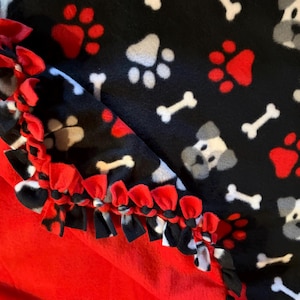 May include: A fleece blanket featuring a black background with a repeating pattern of white dog faces, white bones, and red paw prints. The edge of the blanket is finished with a knotted fringe made from alternating strips of red and black fabric.