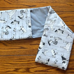 May include: A light gray fabric neck wrap with a pattern of black and white farm animals, including goats, chickens, and dogs. The wrap is folded on a wooden surface, showcasing the patterned fabric and a solid gray interior.