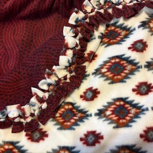 May include: A cozy fleece blanket with a burgundy knit pattern on one side and a white geometric pattern on the other. The edges are finished with a knotted fringe in coordinating colors. A warm and inviting home decor item.
