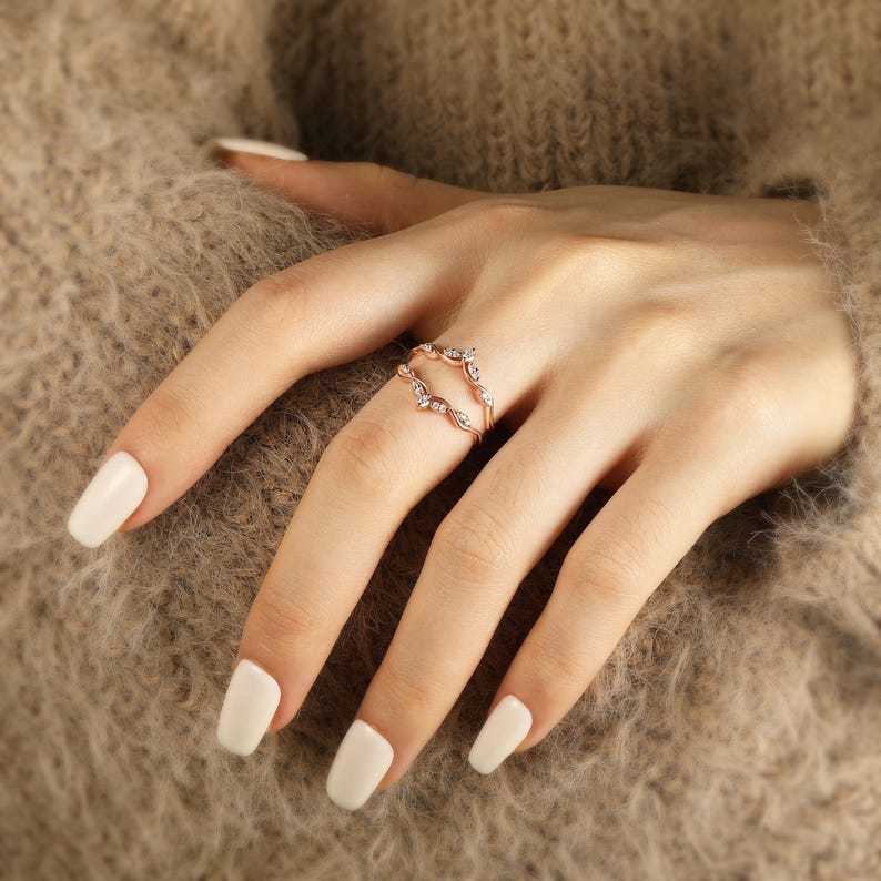 Pu&ograve; includere: Un anello in oro rosa con un design delicato che presenta piccoli diamanti incastonati in un motivo a foglia. L'anello &egrave; indossato su un dito con smalto bianco.