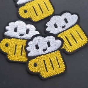 May include: Four embroidered iron-on patches featuring a cartoon beer mug with a white foamy head. The mugs are yellow with black outlines and the foam is white with black eyes and a smiling mouth.