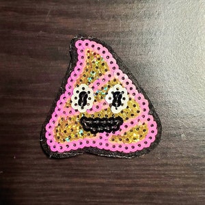 Może przedstawiać: Naszywka z cekinami w kształcie uśmiechniętej emotikony kupy. Emotikona ma złoty środek, różową obwódkę, białe oczy w kształcie kwiatów i czarne, wyszywane usta. Naszywka jest na czarnym filcowym podkładzie.