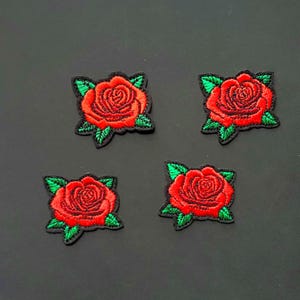 May include: Four embroidered patches featuring a red rose with green leaves on a black background. The roses have detailed stitching and a classic floral design. Each patch is a square shape.