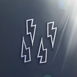 Puede incluir: Cinco parches de hierro en forma de rayo negro y blanco. Los parches miden aproximadamente 5 cm de alto y tienen un contorno blanco.