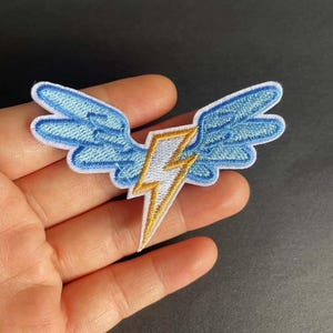 May include: Embroidered patch featuring a blue winged design with a white and gold lightning bolt in the center. The patch has a white border and is likely for clothing or accessories. The design is detailed and colorful.