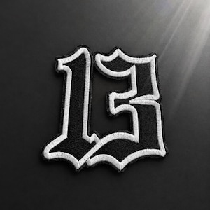 May include: A black and white embroidered patch featuring the number 13 in a stylized gothic font. The design consists of a black numerical character with a thick white border, showcasing detailed stitching throughout the entire surface area.