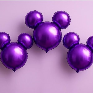 Puede incluir: Tres globos de papel de aluminio morados con forma de Mickey Mouse sobre un fondo lila claro. Los globos tienen un acabado brillante y met&aacute;lico y est&aacute;n dispuestos en fila. Perfecto para una fiesta de cumplea&ntilde;os o evento tem&aacute;tico.