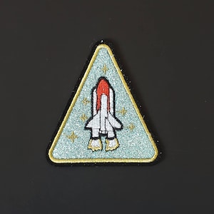 Könnte beinhalten: Ein blauer und goldener dreieckiger Patch mit einem weißen und roten Raumschiff, das darauf gestickt ist. Das Raumschiff ist von kleinen weißen Sternen umgeben.