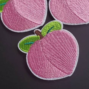 Könnte beinhalten: Drei rosa gestickte Pfirsich-Patches mit grünen Blättern. Die Pfirsiche sind weiß umrandet.