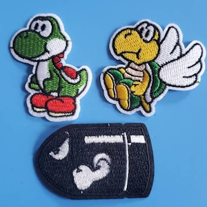 May include: Three embroidered iron-on patches featuring characters from the Super Mario Bros. video game series. The patches are a green Yoshi dinosaur, a yellow Koopa Troopa turtle with wings, and a black and white Bullet Bill.