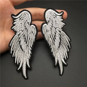 Może przedstawiać: Dwa białe haftowane łaty z skrzydłami anioła z czarnymi obramowaniami. Skrzydła są ozdobione piórami i mają spiczasty koniec.