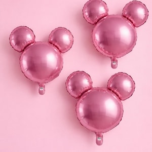 May include: Three pink Mickey Mouse-shaped foil balloons against a pink background. The balloons have a shiny, metallic finish and are arranged in a playful composition, perfect for a party or celebration.