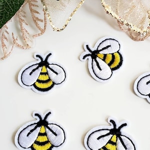 Może przedstawiać: Haftowane naszywki z pszczołami z białymi konturami, czarnymi czułkami i żółto-czarnymi paskami. Naszywki ułożone są na białej powierzchni, z kwiatowymi akcentami w tle.