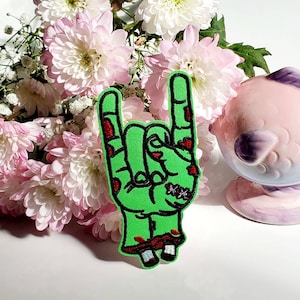 Może przedstawiać: Zielona naszywka z ręką zombie w geście rock and rolla, z czerwonymi akcentami i odsłoniętymi detalami kości. Naszywka znajduje się na białej powierzchni, z różowymi i białymi kwiatami w tle.