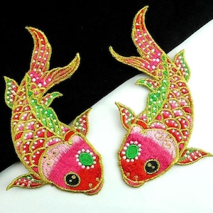May include: Two embroidered koi fish patches. The patches feature a vibrant color palette of red, pink, green, and gold. The fish have detailed scales and fins, with a central eye detail. The patches are likely for decorative purposes, such as embellishing clothing or accessories.