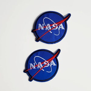 May include: Two blue and white NASA embroidered patches with a red swoosh. The patches are on a white background.
