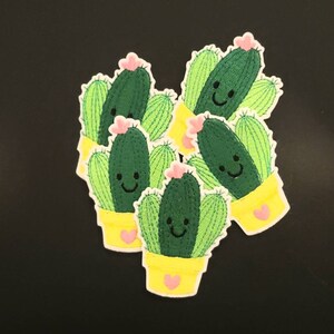 May include: A collection of embroidered cactus patches. Each patch features a smiling green cactus with light green spines, a pink flower, and a yellow pot with a pink heart. The patches are on a black background.