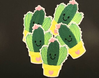 May include: A collection of embroidered cactus patches. Each patch features a smiling green cactus with light green spines, a pink flower, and a yellow pot with a pink heart. The patches are on a black background.