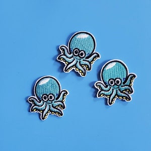 May include: Three blue embroidered octopus patches with white outlines and yellow accents. The octopus have white eyes and a smiling expression.