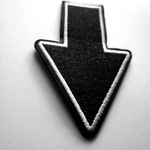 Op de afbeelding: Zwarte pijlpatch met witte rand. De pijl wijst naar beneden. De patch is gemaakt van een textuurstof en staat tegen een witte achtergrond. De pijl is een eenvoudig, grafisch ontwerp.