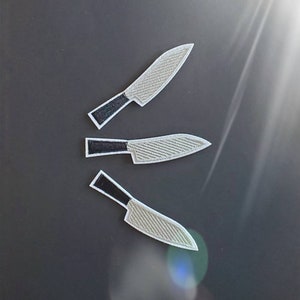 May include: Three embroidered knife patches with silver blades and black handles are arranged on a dark background. The blades have a textured pattern, and the patches are outlined in white. The patches are likely for clothing or accessories.