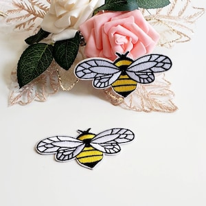 Può includere: Due toppe ricamate a forma di ape con ali bianche e corpi gialli e neri. Le toppe sono su una superficie bianca, con rose rosa e bianche e foglie decorative dorate sullo sfondo.