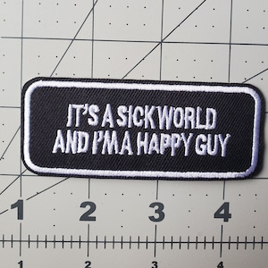 以下が含まれることがあります： 「IT'S A SICK WORLD AND I'MA HAPPY GUY」というテキストが書かれた黒と白の刺繍パッチ。