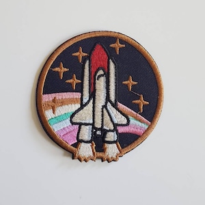 Könnte beinhalten: Ein runder gestickter Aufnäher mit einem Raumschiff, das vor einem dunkelblauen Hintergrund mit goldenen Sternen und einem regenbogenfarbenen Streifen abhebt.