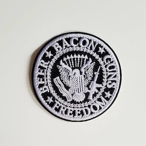 Può includere: Una toppa ricamata in bianco e nero con le parole "Bacon Guns Freedom" che circondano un'aquila che tiene un fucile. L'aquila tiene un fucile nelle sue zampe e ha una stella sul petto.