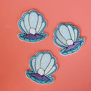 May include: Three embroidered seashell patches. Each patch features a light blue shell with a pearl, outlined in a darker blue. The base of the shell is purple, and the design includes small gold accents.
