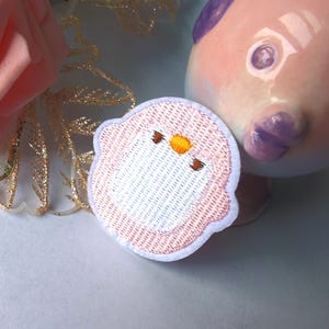 Puede incluir: Parche bordado con un pájaro de dibujos animados con cuerpo blanco, contorno rosa y pico amarillo. El parche tiene un borde blanco. Una figura de pez de cerámica está en el fondo.