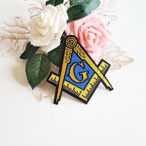 Puede incluir: Un parche bordado en oro y azul con un diseño de compás y cuadrado. El parche presenta una letra "G" en el centro y está rodeado por un borde amarillo.