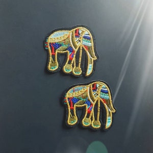 以下が含まれることがあります： 黒い背景に金色の輪郭でカラフルな象が描かれた、アイロン接着式の刺繍パッチ2枚