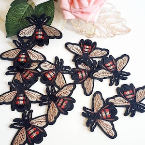 May include: Embroidered bee patches with black outlines, tan wings, and red and black striped bodies are scattered on a white surface. The patches are detailed with intricate stitching. Pink and white flowers are in the background.