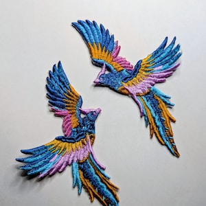 Peut inclure: Deux écussons brodés en forme d'oiseaux aux ailes déployées. Les oiseaux présentent une palette de couleurs vives de bleu, jaune, rose et violet. Les écussons sont probablement destinés à des fins décoratives, comme l'embellissement de vêtements ou d'accessoires.