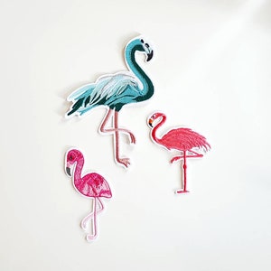 May include: Three embroidered iron-on patches of pink and blue flamingos. The patches are perfect for adding a tropical touch to clothing, bags, or other fabric items.