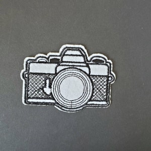 Puede incluir: Un parche bordado en blanco y negro de una cámara vintage. La cámara tiene una lente grande y un cuerpo texturizado.