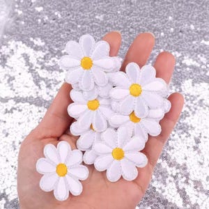 May include: A collection of white daisy flower patches with yellow centers. The patches are made of fabric and have a stitched edge. The daisies are held in a hand against a sparkly silver background. These patches can be used for various craft projects.