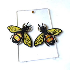 May include: Two yellow and black embroidered bee patches with orange and yellow stripes. The patches are on a clear plastic background.