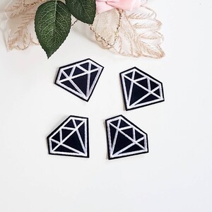 May include: Four diamond-shaped patches with a black background and white embroidered geometric designs. The patches are arranged on a white surface, with a pink rose and green leaves in the background.