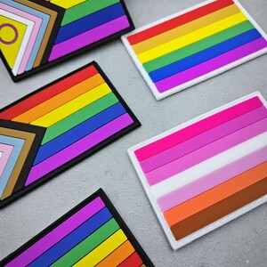 以下が含まれることがあります： さまざまなLGBTQ+プライドフラッグが描かれた、カラフルな長方形のパッチのコレクション。パッチには、レインボーフラッグ、レズビアンフラッグ、プログレスプライドフラッグが含まれており、それぞれ黒い縁と白い縁があります。