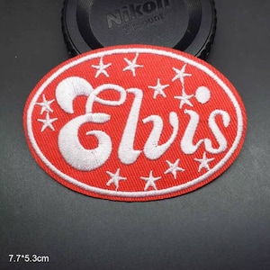 Op de afbeelding: Rode ovale geborduurde patch met witte stiksels. De patch heeft het woord "Elvis" in een gestileerd lettertype omringd door witte sterren.