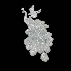 May include: A silver sequined peacock applique with an elaborate tail and floral accents. The decorative embellishment is set against a black background, showcasing the intricate details of the design. Ideal for adding a touch of elegance to garments or crafts.