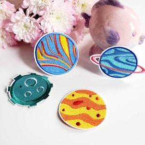 May include: Four embroidered iron-on patches featuring planets in a space theme. The patches are in various colors, including blue, green, orange, and yellow. The planets have different designs, including rings, craters, and stripes.