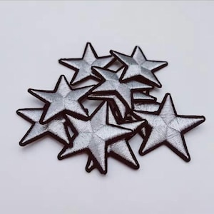 May include: A collection of silver star-shaped embroidered patches. Each patch features a metallic silver thread finish with a distinct five-pointed star design and a dark border outline, arranged in a layered, overlapping cluster formation.