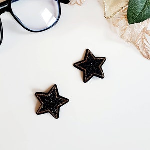 Może przedstawiać: Dwie czarne spinki do włosów w kształcie gwiazdy z złotymi akcentami.