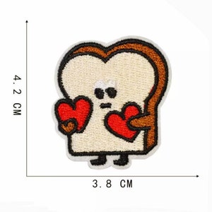 May include: Embroidered patch featuring a cartoon slice of bread with a smiling face and holding two red hearts. The patch is approximately 1.5 inches wide and 1.7 inches tall. The bread has a light tan center and brown crust.
