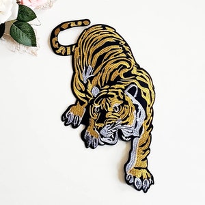 以下が含まれることがあります： 金と黒の刺繍が施されたアイロン接着パッチで、白いディテールが施されています。虎は口を開けて歯を見せて前を向いています。虎は前足を伸ばしたしゃがみ込み姿勢です。