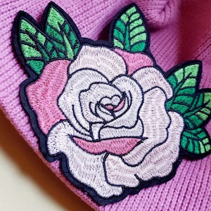 May include: A white and pink embroidered rose with green leaves on a pink knitted fabric.